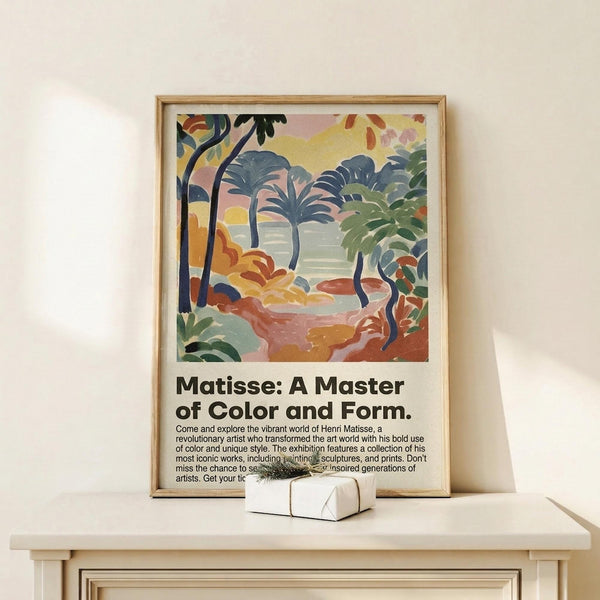 May include: Framed art print featuring a colorful tropical landscape with palm trees, foliage, and water in Matisse's distinctive Fauvist style using vibrant blues, greens, oranges, and pinks. Below the artwork is text reading "Matisse: A Master of Color and Form" with exhibition details. The print sits in a thin wooden frame on a light wood surface next to a small white gift box, displayed against a cream wall.