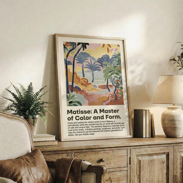 May include: Framed poster featuring a vibrant tropical landscape with palm trees, lush foliage, and warm sunset colors in Matisse's distinctive artistic style, displayed on a rustic wooden sideboard alongside a fern plant, ceramic lamp, and books in a cozy living room setting with neutral tones and natural lighting.