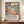 May include: A framed poster featuring a colorful tropical landscape painting with palm trees, foliage, and vibrant blues, greens, yellows, and coral tones in a Fauvist style. The poster includes exhibition text about Matisse below the artwork. The brown wooden frame sits on a wooden surface beside white ceramic vases and books against cream walls with decorative molding.