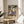 May include: Framed vintage-style travel poster featuring tropical coastal scene with palm trees, sailboats on blue water, Mediterranean buildings, and "MATISSE" text at bottom. Poster sits on round wooden table next to beige upholstered chair with plant shadows visible on white wall behind.