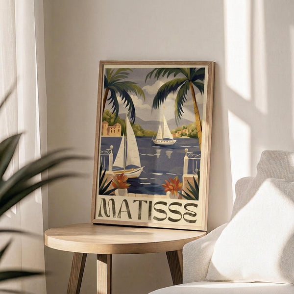 May include: Framed vintage-style travel poster featuring tropical coastal scene with palm trees, sailboats on blue water, Mediterranean buildings, and "MATISSE" text at bottom. Poster sits on round wooden table next to beige upholstered chair with plant shadows visible on white wall behind.