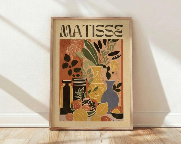 May include: Framed art print in natural wood frame leaning against white wall on light wooden floor. Poster features Henri Matisse-inspired still life with colorful vases, plants, fruits, and vessels in warm earth tones of orange, yellow, green, and blue. Bold "MATISSE" text at top. Artwork shows cut-out style botanical elements including leaves and citrus slices arranged with pottery and bowls in modernist composition.