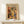 May include: Framed art print in natural wood frame displaying colorful still life with vases, plants, and fruits in Matisse's distinctive cut-out style. Warm earth tones of orange, yellow, green, and blue create a vibrant botanical composition. Print sits on light wood surface beside small wrapped gift with evergreen sprig against cream wall.
