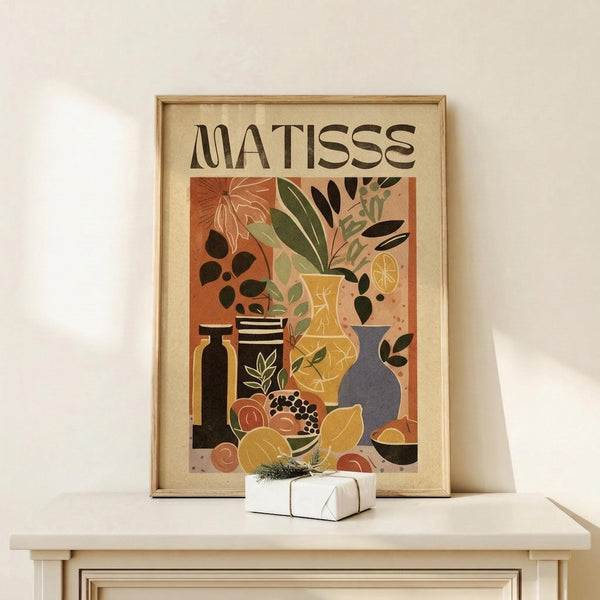 May include: Framed art print in natural wood frame displaying colorful still life with vases, plants, and fruits in Matisse's distinctive cut-out style. Warm earth tones of orange, yellow, green, and blue create a vibrant botanical composition. Print sits on light wood surface beside small wrapped gift with evergreen sprig against cream wall.