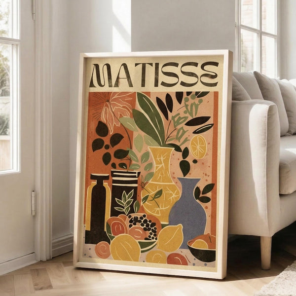 May include: Framed art print in light wood frame leaning against white wall next to beige sofa. Poster features "MATISSE" text above colorful still life with vases, plants, fruits and decorative objects in warm oranges, yellows, greens and blues. Modern living room setting with large windows and natural lighting creates shadows on hardwood floors.