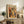 May include: Framed poster with "MATISSE" text and colorful still life artwork featuring vases, plants, and fruits in warm orange, yellow, and green tones. Displayed on rustic wooden sideboard alongside books, ceramic vase, and fern plant in cozy living room setting with beige chair and neutral walls.