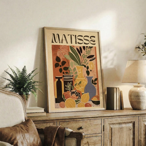 May include: Framed poster with "MATISSE" text and colorful still life artwork featuring vases, plants, and fruits in warm orange, yellow, and green tones. Displayed on rustic wooden sideboard alongside books, ceramic vase, and fern plant in cozy living room setting with beige chair and neutral walls.