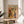 May include: Framed art print featuring a colorful still life composition with potted plants, vases, fruits, and decorative objects in warm earth tones of orange, yellow, green, and blue. The word "MATISSE" appears at the top in bold black letters. The wooden-framed poster sits on a round light wood side table next to a beige upholstered chair, with natural sunlight streaming through white curtains and a plant visible in the corner of the room.