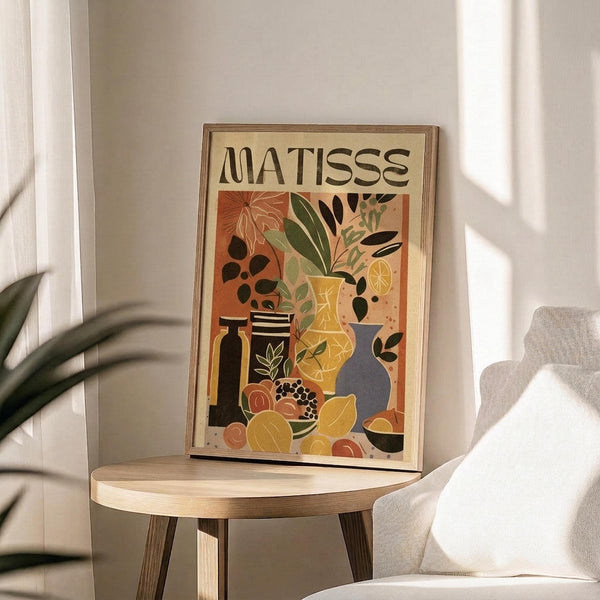 May include: Framed art print featuring a colorful still life composition with potted plants, vases, fruits, and decorative objects in warm earth tones of orange, yellow, green, and blue. The word "MATISSE" appears at the top in bold black letters. The wooden-framed poster sits on a round light wood side table next to a beige upholstered chair, with natural sunlight streaming through white curtains and a plant visible in the corner of the room.