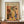 May include: Framed poster featuring Henri Matisse-inspired still life artwork with botanical motifs, ceramic vases, and fruits in warm earth tones of orange, yellow, green, and black. The print displays "MATISSE" text at top and shows various plants, vessels, and organic shapes in a modernist style. Wooden frame sits on credenza beside white ceramic vase and stacked books in neutral-toned room with white walls and crown molding.
