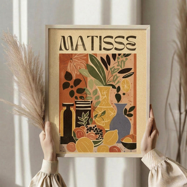 May include: Person holding framed art print featuring Henri Matisse-inspired still life with colorful vases, plants, and fruit in warm orange, yellow, green, and black tones. Light wood frame against neutral background with decorative pampas grass stems visible on left side. Hands wearing cream-colored ruffled sleeves display the vintage-style botanical artwork.