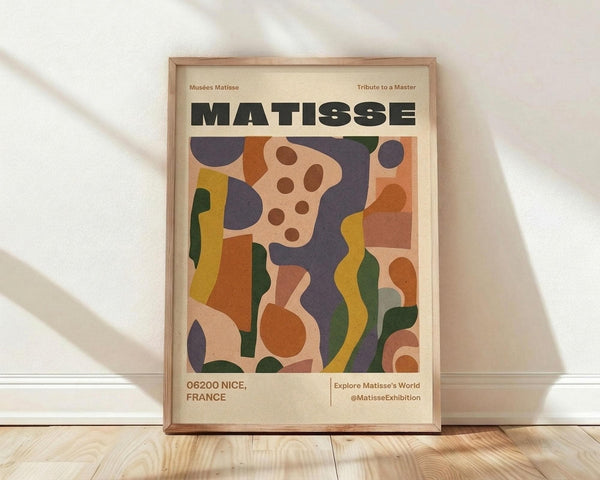 May include: Framed poster print leaning against white wall on wooden floor. Abstract artwork features organic shapes in orange, purple, green, yellow, and pink. Large "MATISSE" text at top, "06200 NICE, FRANCE" at bottom. Natural wood frame, soft window light casting shadows across white textured wall.