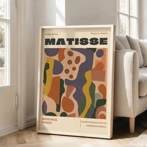 May include: Framed Matisse exhibition poster with colorful abstract cut-out shapes in orange, purple, yellow, and green, displayed against white wall next to beige sofa in bright living room with large windows and natural lighting.