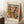 May include: Framed Matisse exhibition poster with colorful abstract cut-out style artwork in orange, purple, green, and yellow organic shapes. Poster leans against white wall on fluffy white rug next to wooden side table with white coffee cup and saucer. Cream-colored blanket visible at right edge.