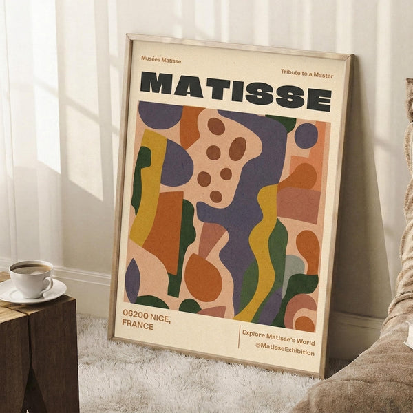 May include: Framed Matisse exhibition poster with colorful abstract cut-out style artwork in orange, purple, green, and yellow organic shapes. Poster leans against white wall on fluffy white rug next to wooden side table with white coffee cup and saucer. Cream-colored blanket visible at right edge.