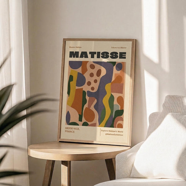 May include: Framed art print featuring abstract colorful shapes in blues, oranges, greens, and yellows with "MATISSE" text at top, displayed on light wood round side table next to beige upholstered chair and plant leaves visible in corner, soft natural lighting creating shadows on neutral wall background.