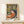 May include: Framed art print featuring a reclining woman with dark hair in orange clothing surrounded by tropical foliage in yellow, green, and coral tones. The poster displays in a light wood frame on a white shelf beside a small wrapped gift with greenery. Soft natural lighting creates gentle shadows on the cream wall behind.