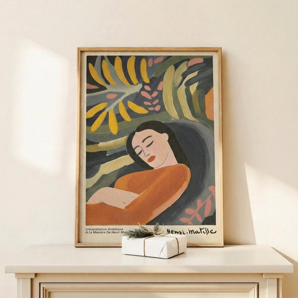 May include: Framed art print featuring a reclining woman with dark hair in orange clothing surrounded by tropical foliage in yellow, green, and coral tones. The poster displays in a light wood frame on a white shelf beside a small wrapped gift with greenery. Soft natural lighting creates gentle shadows on the cream wall behind.