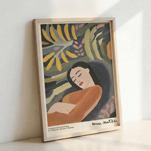 May include: Framed art print showing a reclining woman with dark hair wearing an orange top against a background of tropical foliage in yellow, green, and pink tones. The artwork features a modernist style with simplified forms and muted colors. The piece is displayed in a light wooden frame and positioned against a white wall, with "Henri Matisse" signature visible at bottom right and French text below the image.