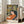 May include: Framed art print leaning against beige sofa in bright living room with tall windows. Print shows woman with long dark hair in orange dress reclining among tropical foliage in green, yellow, pink tones. Matisse-inspired style with bold colors and organic shapes. Light wood frame on hardwood floors.