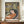 May include: Framed poster print displayed on wooden sideboard featuring a reclining woman with dark hair in orange dress surrounded by colorful tropical foliage in yellow, green, and pink tones. The artwork shows Matisse-inspired style with bold, simplified forms and vibrant colors against a dark background. White ceramic vase sits beside the brown-framed print in an elegant room with cream walls and white molding.