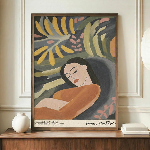 May include: Framed poster print displayed on wooden sideboard featuring a reclining woman with dark hair in orange dress surrounded by colorful tropical foliage in yellow, green, and pink tones. The artwork shows Matisse-inspired style with bold, simplified forms and vibrant colors against a dark background. White ceramic vase sits beside the brown-framed print in an elegant room with cream walls and white molding.