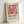 May include: Framed poster print leaning against white wall showing colorful Matisse-style interior scene with red window frames, orange flowers in vase, green plants, and blue water view. Features exhibition text "Matisse: A Master of Color and Form" with descriptive paragraph below artwork. Light wood frame on white surface with soft lighting.