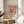 May include: A framed poster featuring a vibrant Matisse-style interior scene with red curtains, blue window frame, colorful flowers, and green plants, displayed on a light wood round table next to a beige upholstered chair. Natural sunlight streams across white walls, with a plant's shadow visible in the corner. The artwork shows bold colors and flat shapes typical of Matisse's fauve style.