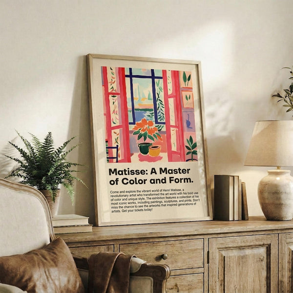 May include: Framed Matisse exhibition poster featuring colorful window scene with red curtains, potted plants, and view to water, displayed on rustic wooden sideboard alongside fern plant, books, and ceramic vases in warm-toned living room setting with beige sofa and neutral walls.