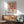 May include: Framed art print of a Matisse-style interior scene with red window frames, colorful plants, and ocean view, displayed above a mid-century modern wooden sideboard with ceramic lamp, next to a sage green upholstered armchair with walnut frame in a sunlit room with white walls.