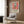 May include: Matisse exhibition poster in light wooden frame mounted on wood-paneled wall above beige upholstered chair with geometric pillow. Poster features colorful interior scene with red window frames, plants, and vessels in Matisse's distinctive style. Modern reading nook with brass floor lamp, side table with open book, and hardwood floors visible.
