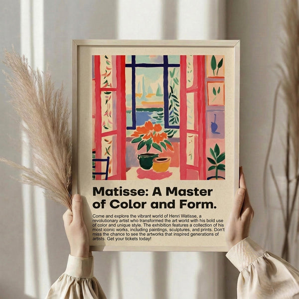 May include: Person in beige ruffled sleeve top holding framed colorful Matisse-style poster showing interior scene with red open window, potted plants, and ocean view. Dried pampas grass visible beside frame against neutral curtained background. Poster features vibrant blues, reds, greens in fauve art style with exhibition text below artwork.