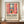 May include: A framed poster featuring a Matisse-style colorful interior scene with red window frames, potted flowers, and view of boats on water, displayed on a wooden console table next to a white ceramic vase and stacked books in a bright room with white walls and crown molding.