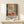 May include: Framed art poster in light wood frame showing Matisse-style colorful interior scene with open window, plants, and geometric shapes in blues, oranges, and greens. Text reads "Discover the Art of Henri Matisse" with exhibition details. Displayed on light wood surface with small wrapped gift box beside it against cream wall.