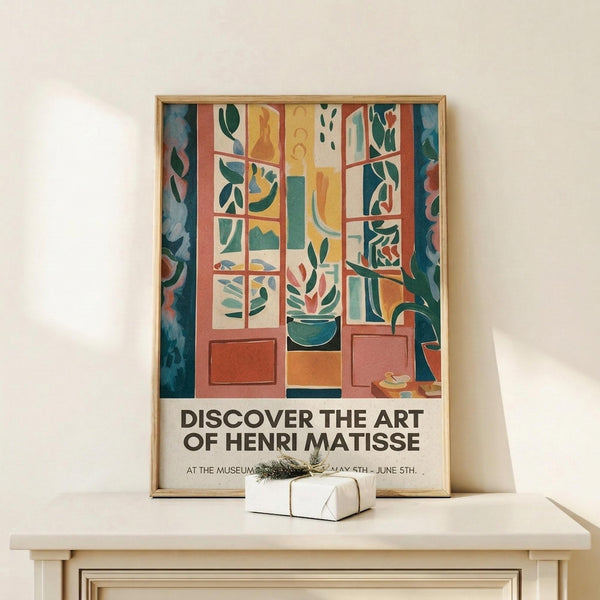 May include: Framed art poster in light wood frame showing Matisse-style colorful interior scene with open window, plants, and geometric shapes in blues, oranges, and greens. Text reads "Discover the Art of Henri Matisse" with exhibition details. Displayed on light wood surface with small wrapped gift box beside it against cream wall.