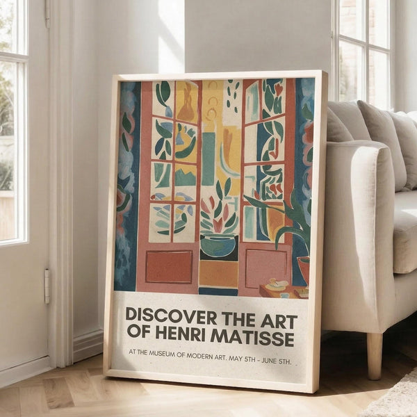 May include: Colorful Henri Matisse exhibition poster in light wood frame leaning against white wall next to beige sofa. Artwork shows vibrant window scene with geometric panels in blues, oranges, yellows and greens, featuring botanical motifs and interior elements. Bold black text reads "DISCOVER THE ART OF HENRI MATISSE" with museum details below. Bright natural light streams through tall windows in modern living room setting.