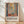 May include: Framed art poster leaning against white wall on light wood floor with plant shadow overlay. Artwork shows colorful interior scene with window view, featuring green foliage, potted plants, and warm earth tones in geometric composition. Natural wood frame, museum exhibition style text below image.