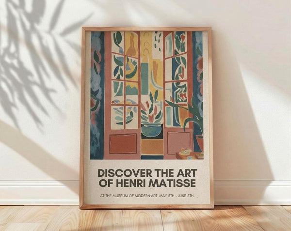 May include: Framed art poster leaning against white wall on light wood floor with plant shadow overlay. Artwork shows colorful interior scene with window view, featuring green foliage, potted plants, and warm earth tones in geometric composition. Natural wood frame, museum exhibition style text below image.