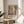 May include: Framed art print displayed on round wooden side table next to beige upholstered chair. Poster features colorful abstract interior scene with window, plants, and geometric shapes in blues, oranges, and greens. Text reads "Discover the Art of Henri Matisse" with museum exhibition details. Natural lighting creates soft shadows across neutral-toned room setting.