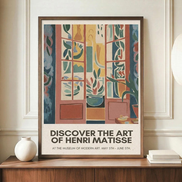 May include: Framed exhibition poster featuring Henri Matisse's colorful fauve-style artwork depicting an open window view with green foliage, potted plants, and geometric interior elements in vibrant blues, oranges, and greens. Text reads "Discover the Art of Henri Matisse" and "At the Museum of Modern Art, May 5th - June 5th." Poster displayed on wooden credenza beside white ceramic vase and stacked books in elegant room with cream walls and white crown molding.