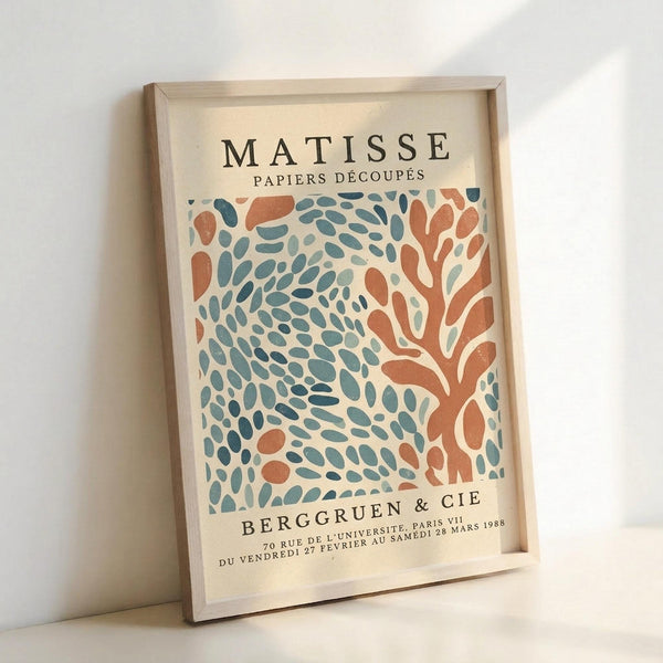 May include: Framed vintage exhibition poster featuring Matisse's cut-out paper art style with organic coral and teal blue shapes resembling leaves and coral branches on cream background. Light wood frame displayed against white wall, casting soft shadows. French text reads "MATISSE PAPIERS DÉCOUPÉS" at top and "BERGGRUEN & CIE" with exhibition details at bottom in black typography.