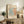 May include: Framed Matisse exhibition poster with coral and blue-green cut-out leaf design displayed on rustic wooden sideboard. Room scene shows warm neutral walls, fern plant, ceramic vase, stacked books, and brown leather furniture. Poster features vintage typography and organic flowing shapes in earthy color palette.