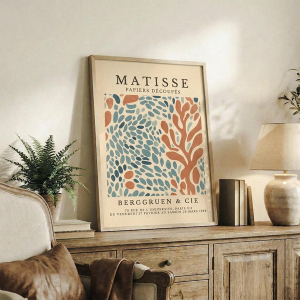 May include: Framed Matisse exhibition poster with coral and blue-green cut-out leaf design displayed on rustic wooden sideboard. Room scene shows warm neutral walls, fern plant, ceramic vase, stacked books, and brown leather furniture. Poster features vintage typography and organic flowing shapes in earthy color palette.