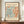 May include: Framed vintage-style exhibition poster featuring abstract cut-paper design in coral orange and teal blue organic shapes on cream background, displayed on wooden sideboard with white ribbed ceramic vase and books, against neutral wall with white molding.