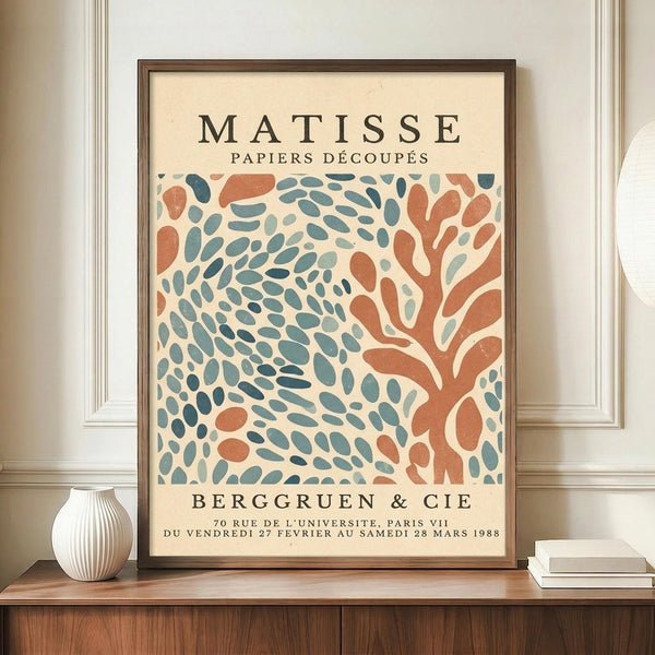 May include: Framed vintage-style exhibition poster featuring abstract cut-paper design in coral orange and teal blue organic shapes on cream background, displayed on wooden sideboard with white ribbed ceramic vase and books, against neutral wall with white molding.