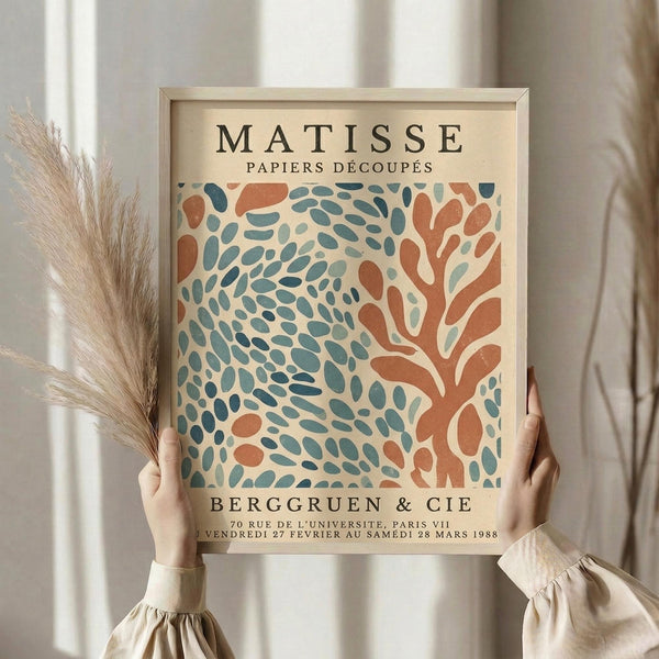 May include: Person in cream blouse holding framed Matisse exhibition poster featuring abstract coral and teal organic shapes on cream background, displayed with dried pampas grass against white curtained window in soft natural lighting