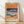 May include: Framed art print featuring abstract landscape with layered horizontal bands in warm oranges, deep blues, teal waves with white foam, and purple-pink sections, displayed in light wood frame against white wall on wooden floor with natural lighting creating diagonal shadows