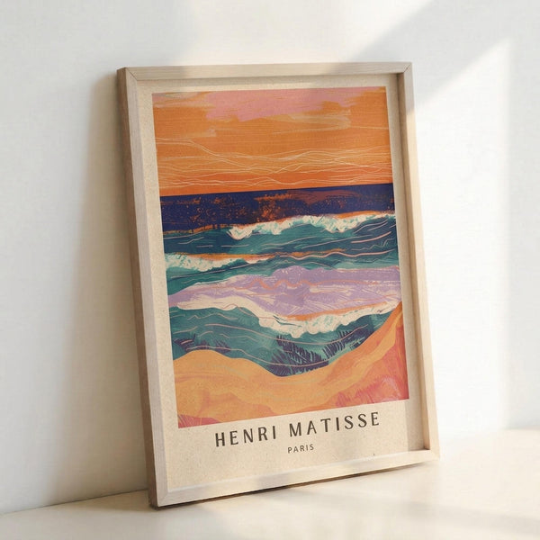 May include: Framed art print leaning against white wall showing abstract seascape with layered horizontal bands in orange sunset sky, deep blue water, teal and green waves with white foam, and sandy beach foreground. Natural wood frame on light wooden surface.