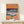 May include: Framed art print featuring an abstract seascape with vibrant orange and pink sky, deep blue ocean with white foam waves, and coral-colored sandy shore. The colorful poster displays flowing, layered brushstrokes in a modern artistic style with "HENRI MATISSE" text at bottom. Light wood frame rests on white wooden surface next to small wrapped gift box with evergreen sprig.