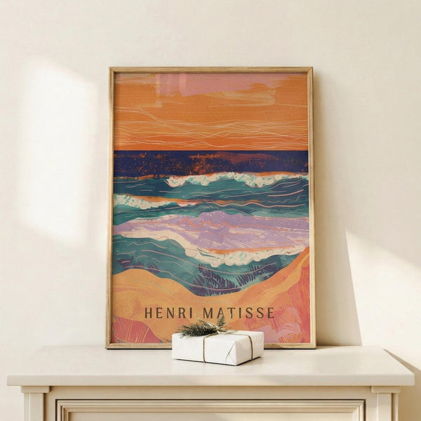May include: Framed art print featuring an abstract seascape with vibrant orange and pink sky, deep blue ocean with white foam waves, and coral-colored sandy shore. The colorful poster displays flowing, layered brushstrokes in a modern artistic style with "HENRI MATISSE" text at bottom. Light wood frame rests on white wooden surface next to small wrapped gift box with evergreen sprig.
